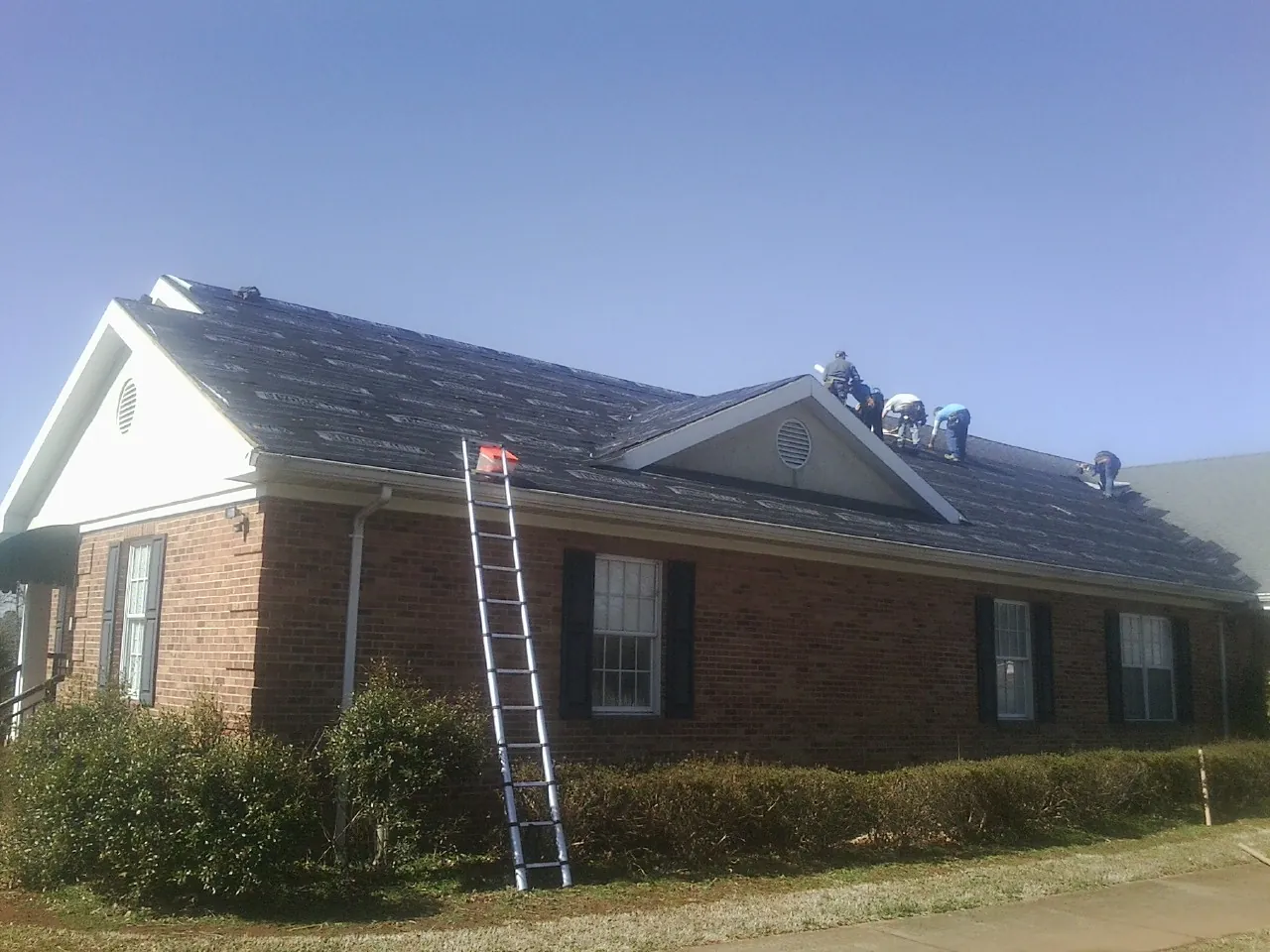 Crew tearing shingles off a brick ranch roof with a ladder set — Emergency Roof Repair work by our crew in Corning